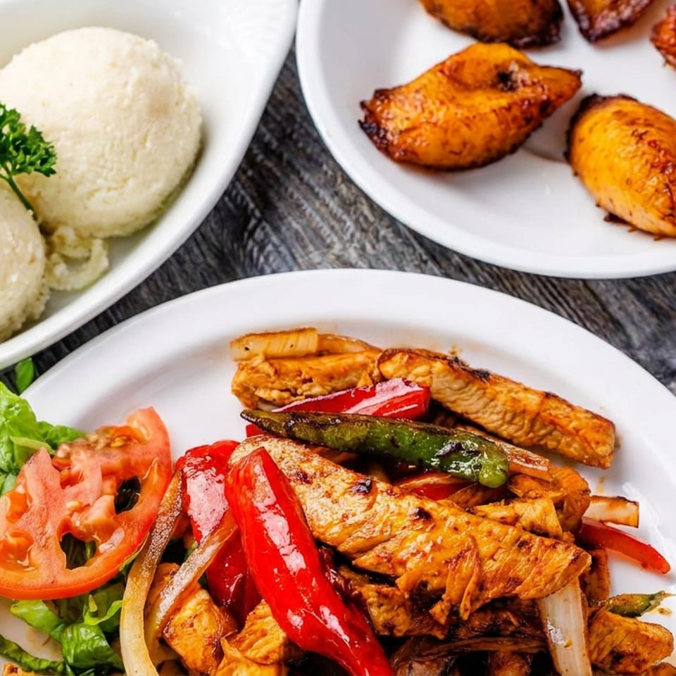 Plates of grilled chicken with peppers and vegetables, white rice dumplings, and roasted plantains on a wooden table.