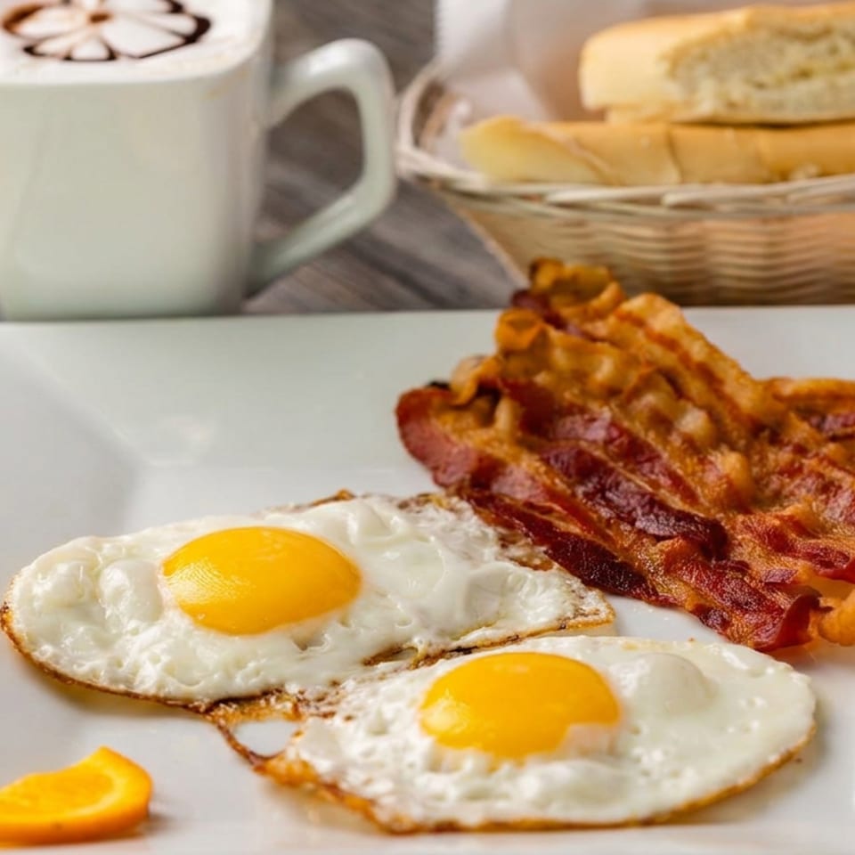 Breakfast plate with two fried eggs, crispy bacon, toast basket, and a cup of coffee on a wooden table.