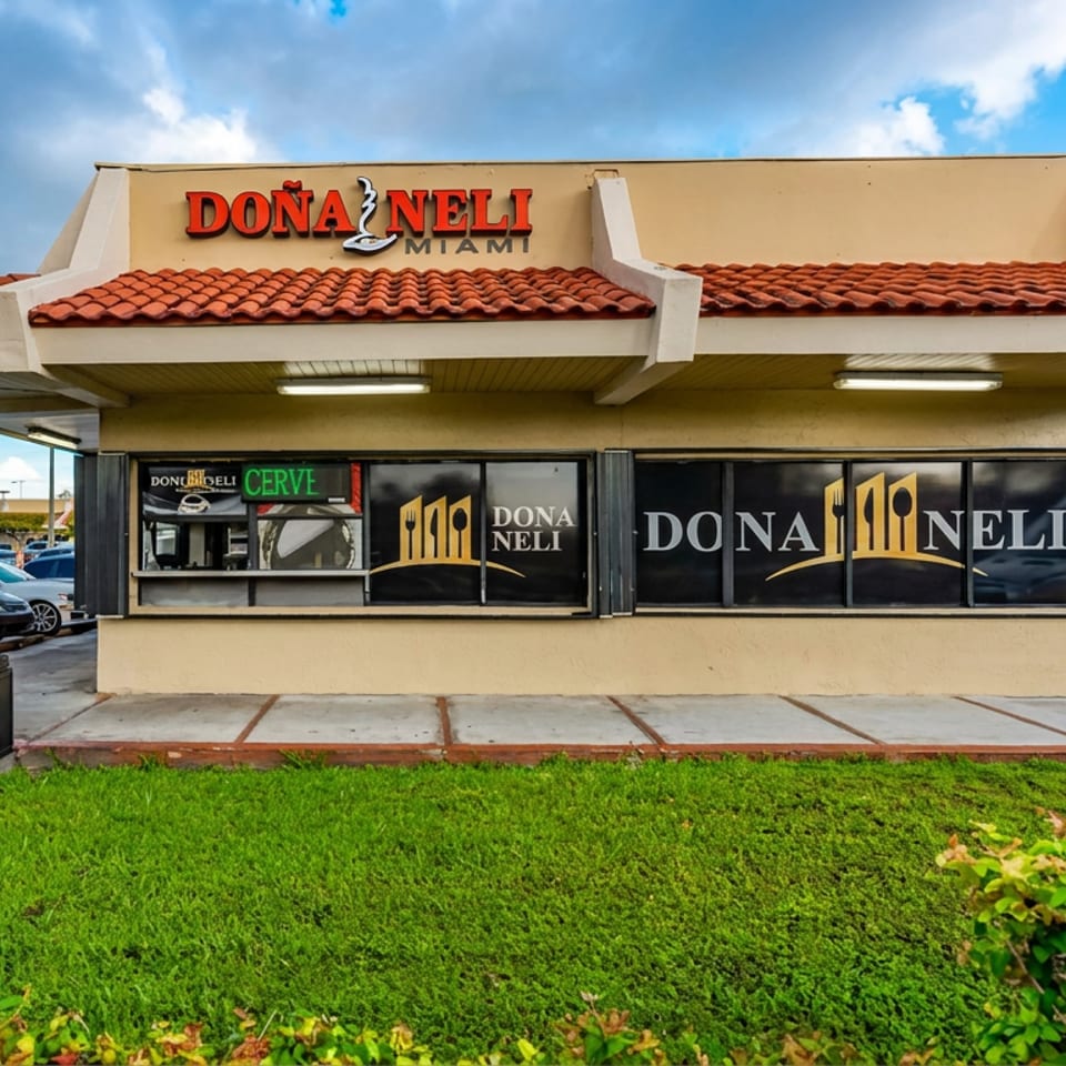 Exterior of Dona Neli Miami restaurant with red tile roof, branded windows, palm trees, landscaping, and parked cars.