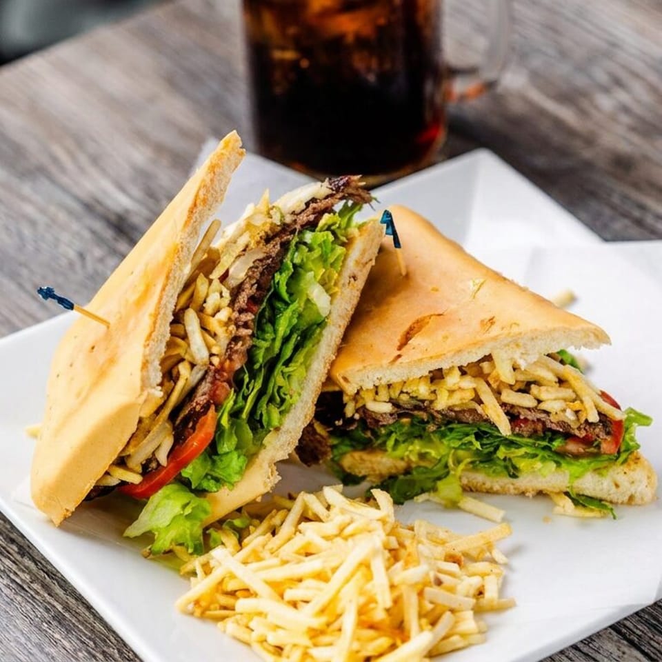 Two toasted sandwiches filled with meat, lettuce, tomatoes, and crispy potato sticks on a white plate, with extra sticks and a soda.