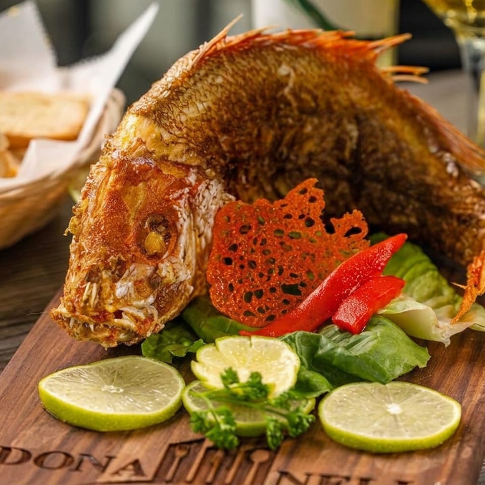 Whole fried fish served on a wooden board with lettuce, peppers, lime slices, bread basket, and white wine.