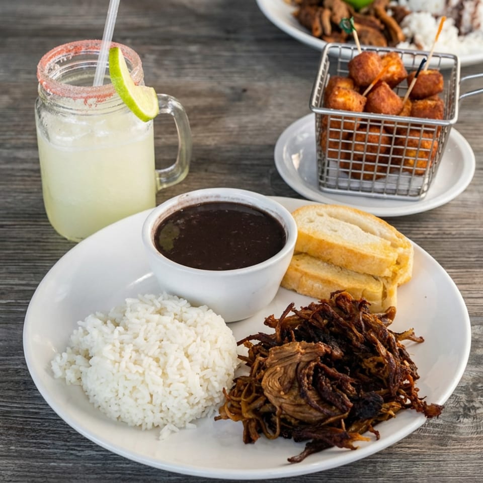 Latin American meal with white rice, shredded beef, beans, fried plantains, dipping sauce, limeade, and fried appetizers on a wooden table.