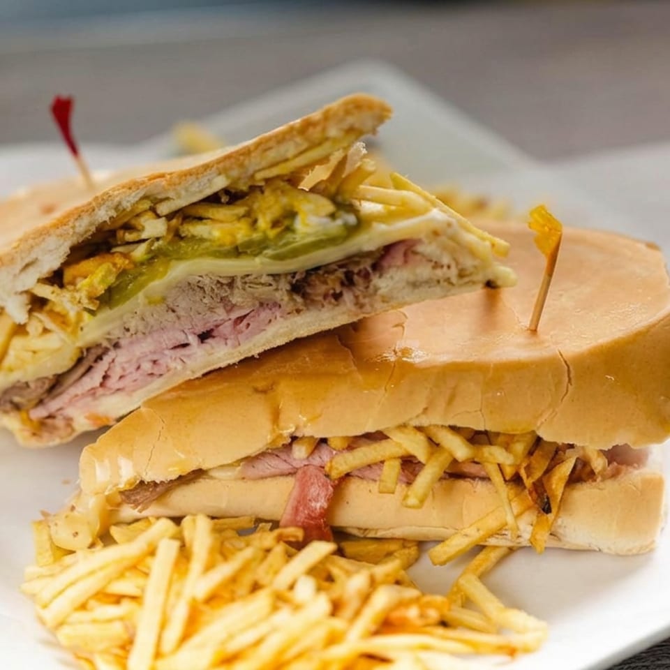 Two toasted deli sandwiches filled with sliced meat, cheese, lettuce, and crispy potato sticks, served with fries on a white plate.