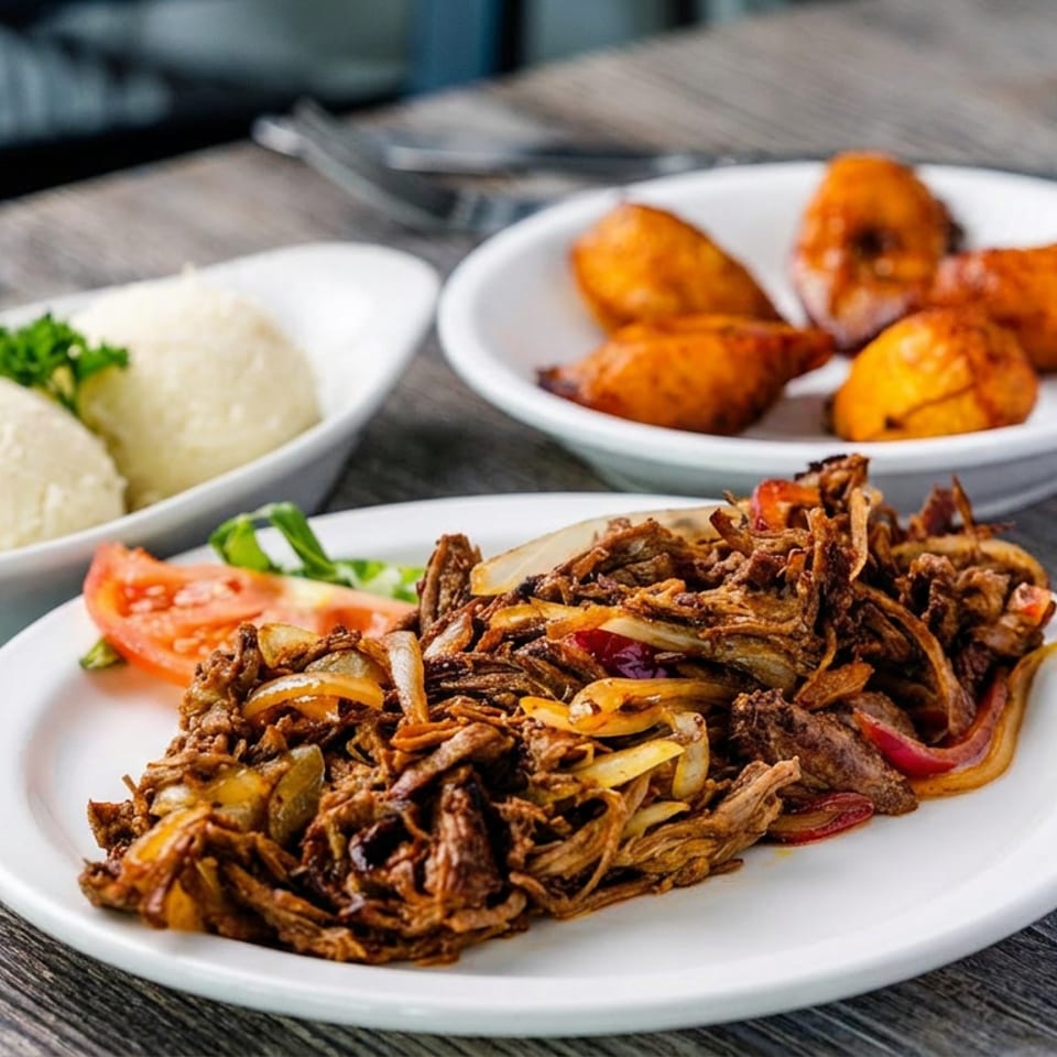 Plates of shredded spiced meat with vegetables, white rice, and golden fried plantains on a wooden restaurant table.