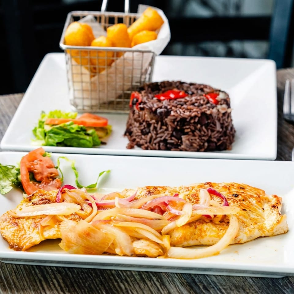 Two plated meals on a wooden table: grilled fish with onions, black beans and rice, salad, and fries.