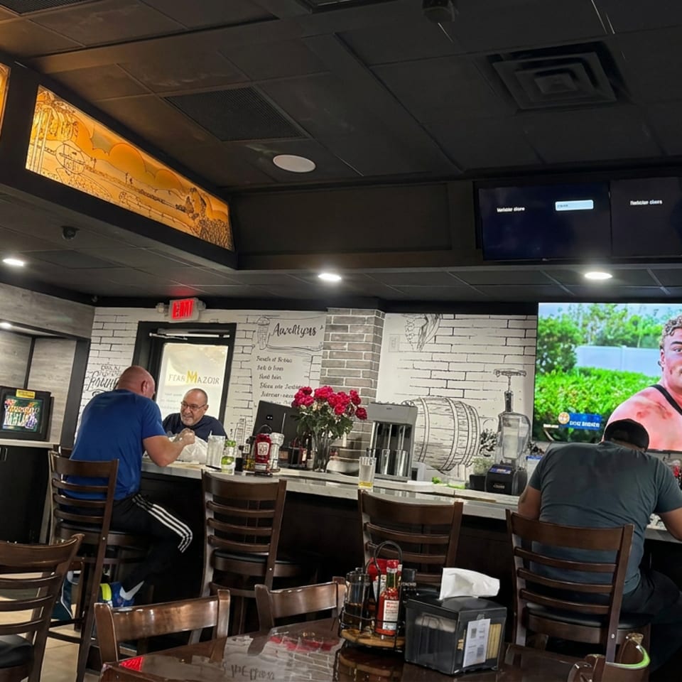 Busy restaurant bar with seated patrons, illuminated mural panels, wine-themed decor, and televisions showing a sporting event.